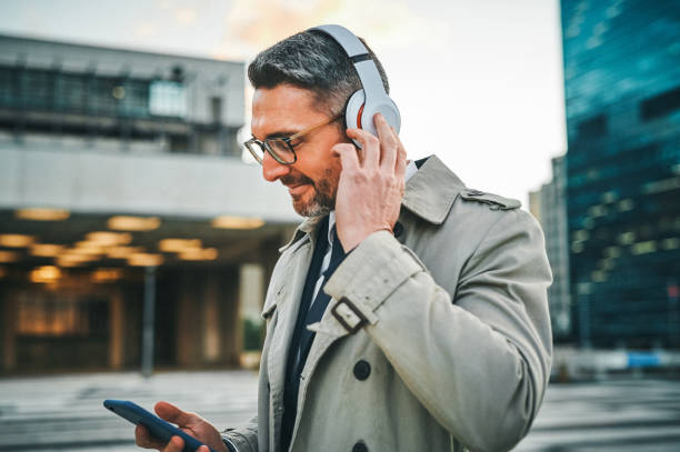 Shot of a mature businessman wearing headphones and using a cellphone in the city Catching up with podcasts during his morning commute podcast stock pictures, royalty-free photos & images