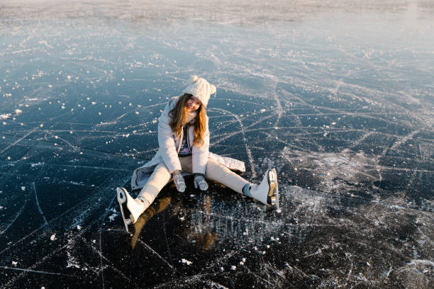 A young woman is sitting in skates on the ice of a frozen lake. Sunny day, atmosphere of fun winter activities. stock photo