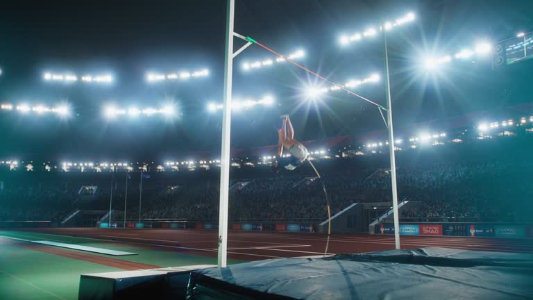 Pole Vault Jumping Championship: Professional Male Athlete Running with Pole Successfully Jumping over Bar and Landing on His Feet, Celebrates Record Setting Victory with Stadium Crowd Cheering