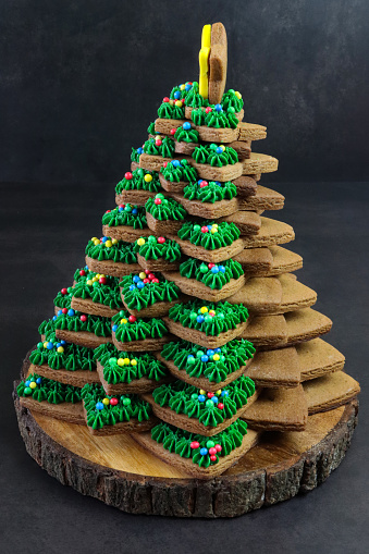 Stock photo showing close-up view of wooden cake stand containing a stack of star shaped gingerbread biscuits, partially decorated with green butter icing and multi-coloured candy coated sweets, forming a Christmas tree. Home baking concept.