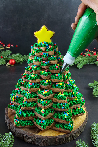 Stock photo showing close-up view of wooden cake stand containing a stack of star shaped gingerbread biscuits, decorated with green butter icing and multi-coloured candy coated sweets, forming a Christmas tree, surrounded by spruce needles and red berries. Home baking concept.