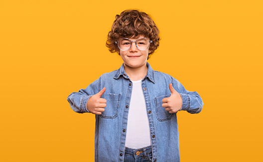 Optimistic curly haired boy in denim clothes and glasses showing thumbs up and looking at camera isolated against bright yellow background