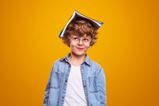 Curious pupil with textbook on head Funny clever schoolboy with open textbook on head smiling and looking away while doing homework against yellow background small boys stock pictures, royalty-free photos & images