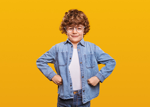 Proud boy in glasses and denim jacket flexing biceps and smiling for camera while showing strength against yellow background