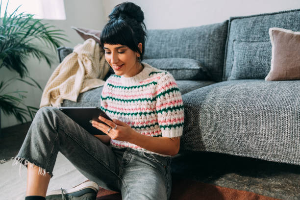 une jeune femme assise par terre et regardant un podcast sur sa tablette - simplicity using computer women computer equipment photos et images de collection