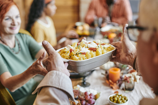 Hand of mature man passing bowl with baked pasta to his wife while both sitting by festive table against their family