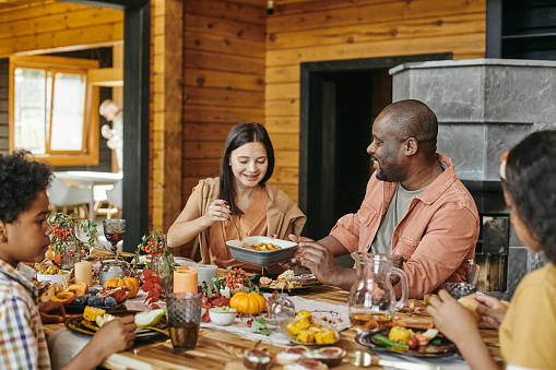 Happy young woman passing her husband bowl with baked potatoes during family dinner by festive table
