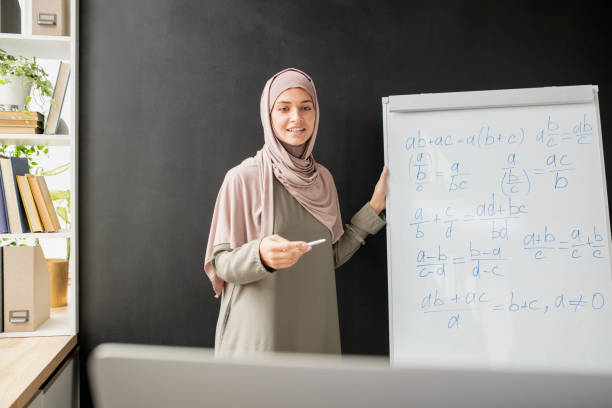 istock Happy young teacher in hijab standing by whiteboard with algebraic formula and equations 1357574792