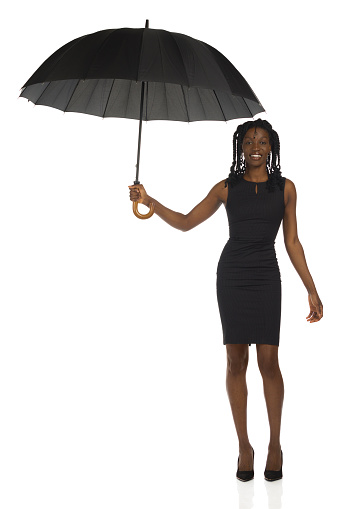 Elegant african woman in mini dress and high heels is standing and holding big open umbrella. Front view. Full length on white background.