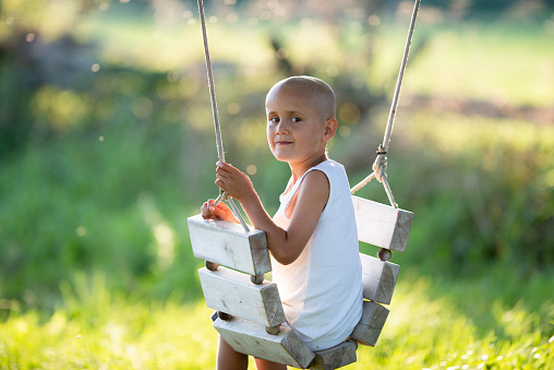 A young boy with cancer is seen sitting on a wooden swing outside at he takes a brief pause form his treatments to enjoy the fresh air.  He is dressed casually in a white tank top and has his head shaved as he looks over his shoulder with a smile.
