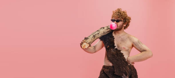 abgeschnittenes porträt eines künstlerischen mannes im charakter eines neandertalers, der tierhauttuch trägt und kaugummi isoliert über rosa hintergrund isst. flugblatt - ethnisches erscheinungsbild stock-fotos und bilder