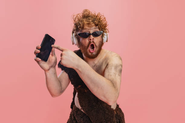 porträt eines mannes im charakter eines neandertalers in kopfhörern, der auf das telefon zeigt, mit schockiertem ausdruck isoliert über rosa hintergrund - ethnisches erscheinungsbild stock-fotos und bilder