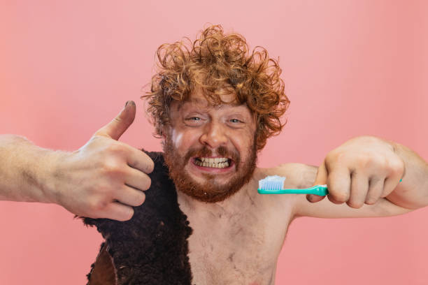 beschnittenes porträt eines lustigen mannes im charakter eines neandertalers, der zähneputzt, mit zahnpasta, die über rosa hintergrund isoliert ist. ähnliche geste anzeigen - ethnisches erscheinungsbild stock-fotos und bilder