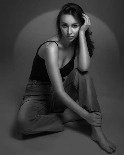 Photo of Fashion model sitting on a floor in a jeans and black undershirt barefoot on studio gray background