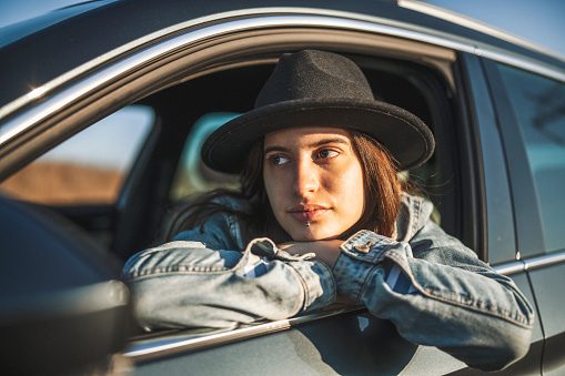 Portrait of young woman with hat leaning on car window and looking away