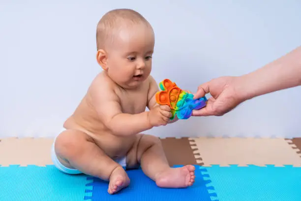 Photo of The father's hand gives the newborn a modern pop-it toy. Child in the playroom on the floor made of soft puzzles
