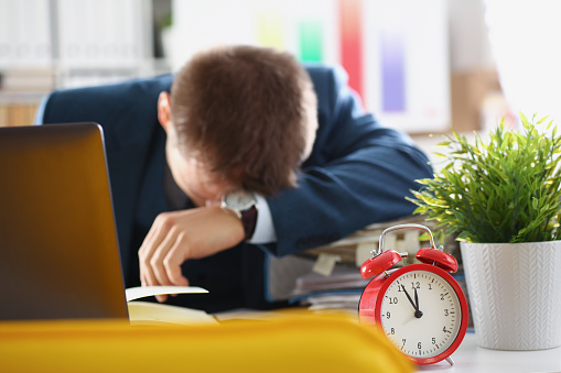 A man sleeps in the workplace, an alarm clock in the foreground. Work fatigue, deadline. Emotional burnout, stress