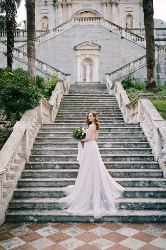 Bride in a long dress stands half-wrapped on stone steps near an old building. High quality photo