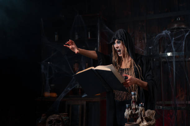 istock Halloween concept. Witch dressed black hood with dreadlocks standing dark dungeon room use magic book for conjuring magic spell 1357618150