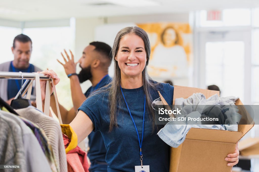 Mid adult female clothing drive organizer poses for photo Busy mid adult female clothing drive organizer smiles for camera.  She is holding a box of donated clothes and stands by a rack of hanging clothes.  Two male volunteers argue in the background. Volunteer Stock Photo