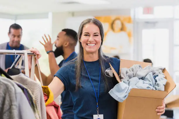 Busy mid adult female clothing drive organizer smiles for camera.  She is holding a box of donated clothes and stands by a rack of hanging clothes.  Two male volunteers argue in the background.