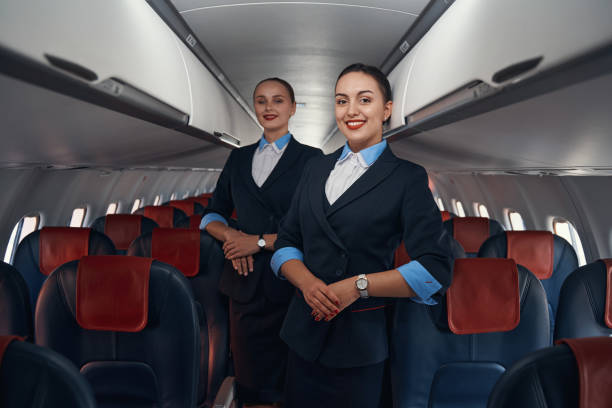 Cabin crew members posing for photo inside plane Two airline hostesses in uniforms standing on the aisle of airplane cabin with smiles on faces and folded hands crew stock pictures, royalty-free photos & images