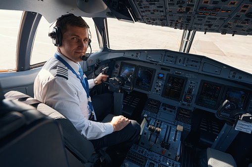 Male pilot sitting at control wheel of passenger airplane and turning back while staring at camera