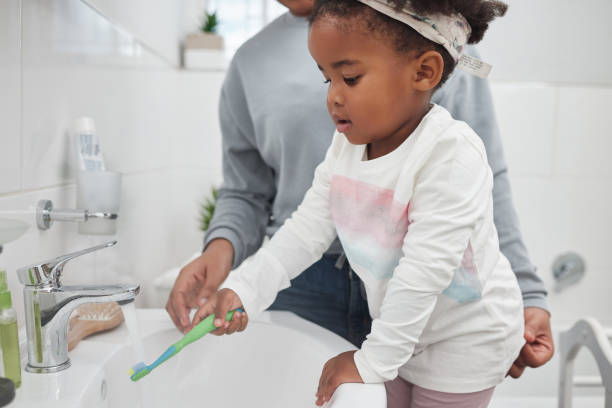 tiro de uma mãe ajudando sua filha pequena enxaguar sua escova de dentes em uma torneira no banheiro em casa - bathroom black faucet - fotografias e filmes do acervo