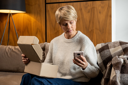 Middle-aged woman sitting on the sofa opens the box, checks the parcel using a smartphone.