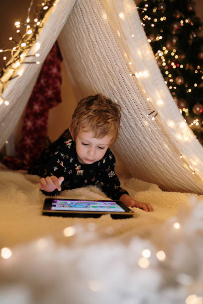 Little boy using digital tablet laying down under tent made from bed sheet stock photo
