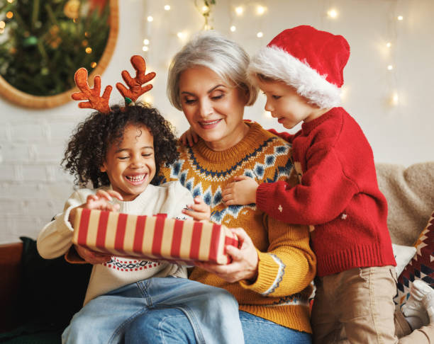 dwóch chłopców różnych ras daje prezent noworoczny babci, siedząc w pobliżu drzewa bożego narodzenia - grandmother giving gift child zdjęcia i obrazy z banku zdjęć