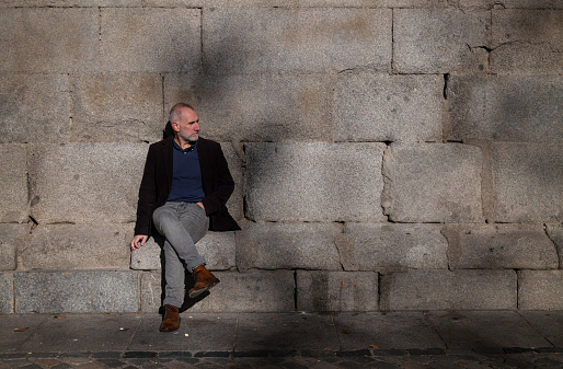 Adult man in suit sitting against stone wall with sunlight and shadow. Shot in Madrid, Spain