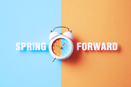 Spring forward reads next to a white alarm clock on blue and salmon background. Horizontal composition with copy space. Spring forward concept.
