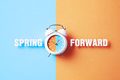 Spring Forward Reads Next To A White Alarm Clock On Blue And Salmon Background