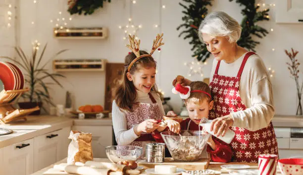 Happy family elderly grandmother and two little kids make Christmas homemade cookies together, standing behind table in kitchen decorated for xmas, children cooking with grandma during winter holidays