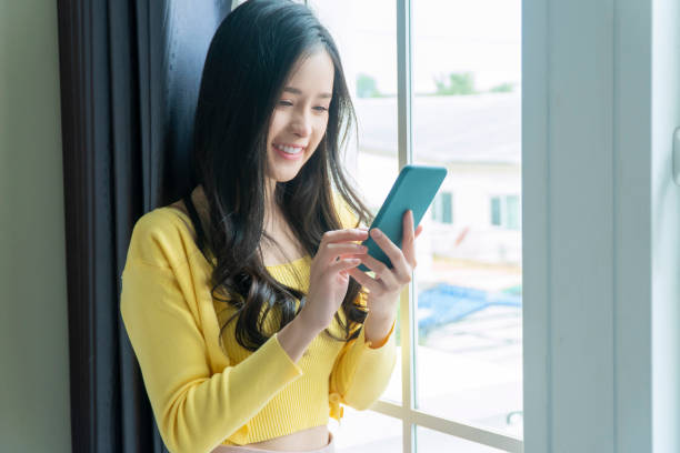 istock Cheerful young woman using smartphone check mail or social network in own hands while standing beside window at home. Lifestyle technology communication concept. 1357647436