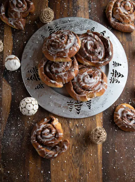 Fresh and homemade baked cinnamon rolls for christmas season served on a silver tray with decoration on rustic and wooden background. Closeup and overhead view
