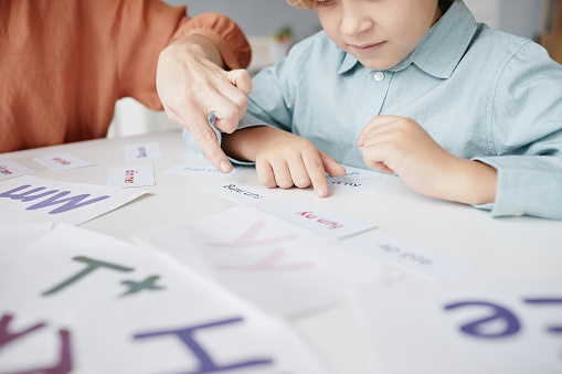 Schoolboy sitting at the table and learning to read English words together with teacher who pointing at text