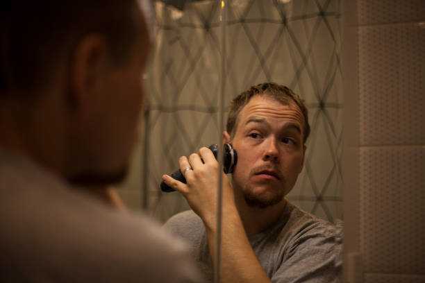 young married man shaves early in the morning in the bathroom and sees his reflection in the mirror. - shaving men electric razor reflection imagens e fotografias de stock