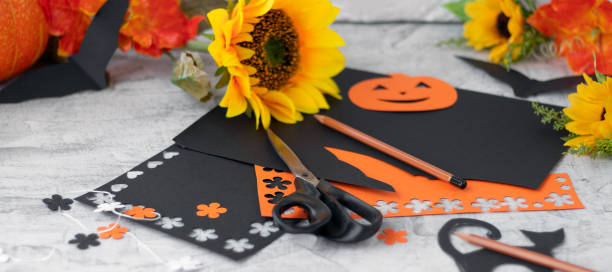 banner with black and orange paper and scissors on a background of autumn flowers and leaves. Preparing for Halloween. making paper garlands at home. stock photo
