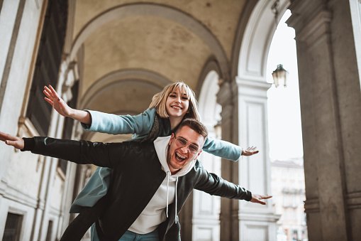 Smiling Male Carrying Girlfriend During Flying Piggyback Ride Through Architectural Arch