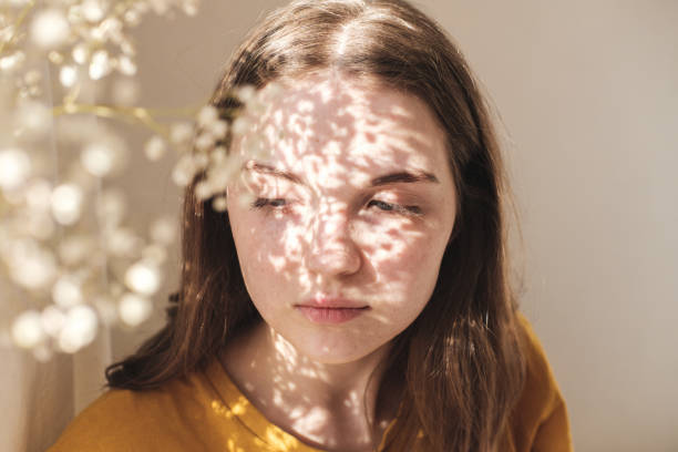 Portrait of young beautiful woman by the window with shadow from flowers on her face. Morning spring aesthetics. stock photo