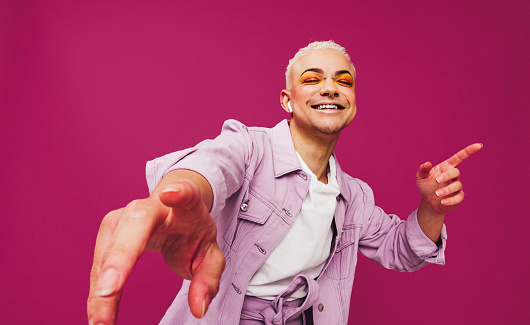 That's my favourite song. Happy young man dancing to his favourite music while wearing wireless earphones. Queer man smiling cheerfully while standing against a purple background.