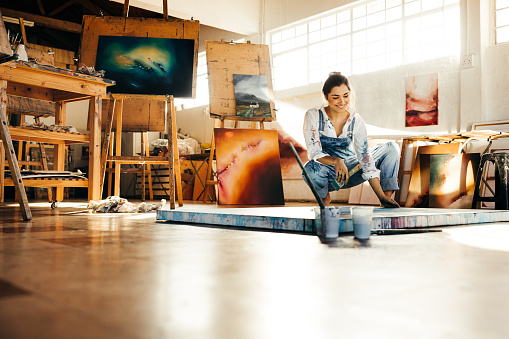 Happy female painter making a blue painting on a canvas. Creative young woman smiling while looking at her painting on the floor. Cheerful young artist working on a new project in her atelier.