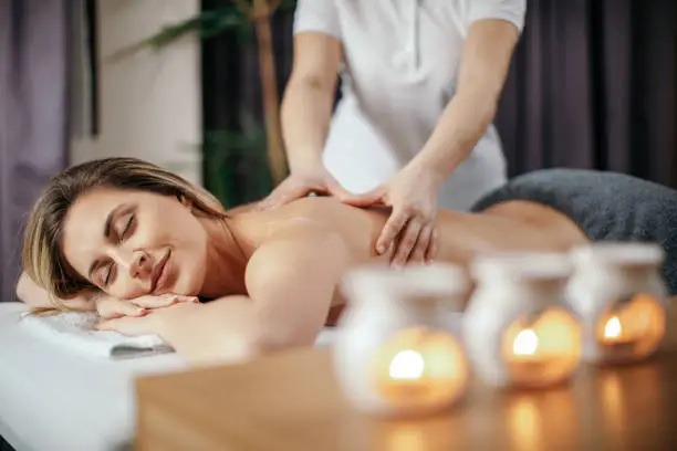 Beautiful young woman receiving relaxation back massage in spa salon. She has a nice healthy skin and looks beautiful and very attractive. Candles with ethereal oil are on the desk in front of her