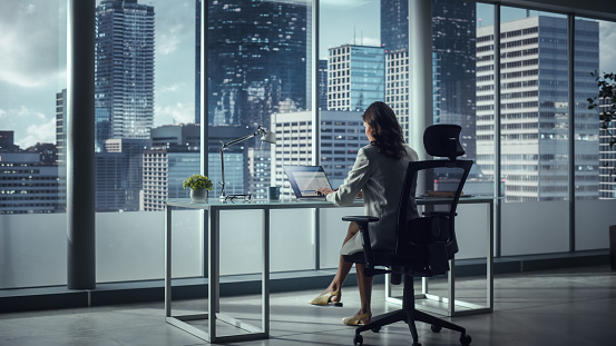 Successful Caucasian Businesswoman Sitting at Her Desk Working on Laptop Computer in Big City Office. Confident Corporation CEO Plan Investment Strategy for Disruptive e-Commerce Startup. Back View