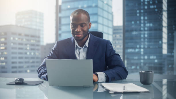 istock Successful Black Businessman in Tailored Suit Working on Laptop Computer on Top Floor Office Overlooking Big City. Successful Software Engineer does Data Analysis for His e-Commerce Startup Investment 1357661139