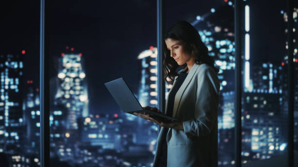 bureau moderne de la grande ville la nuit: jeune femme d’affaires prospère debout et utilisant un ordinateur portable. belle femme entrepreneur numérique pensant à la stratégie d’investissement pour le projet de commerce électronique. - business office business person window photos et images de collection