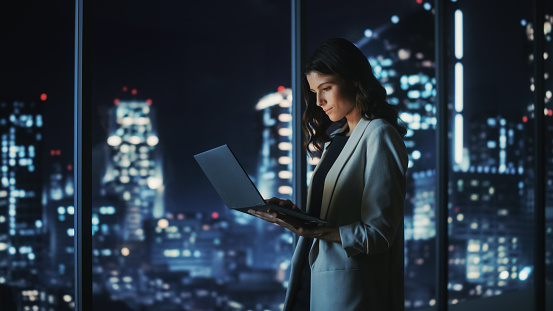 istock Big City Modern Office at Night: Successful Young Businesswoman Standing and Using Laptop. Beautiful Female Digital Entrepreneur Thinking of Investment Strategy for e-Commerce Project. 1357661160