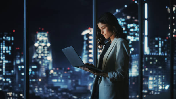 Big City Modern Office at Night: Successful Young Businesswoman Standing and Using Laptop. Beautiful Female Digital Entrepreneur Thinking of Investment Strategy for e-Commerce Project. Big City Modern Office at Night: Successful Young Businesswoman Standing and Using Laptop. Beautiful Female Digital Entrepreneur Thinking of Investment Strategy for e-Commerce Project. digital transformation stock pictures, royalty-free photos & images